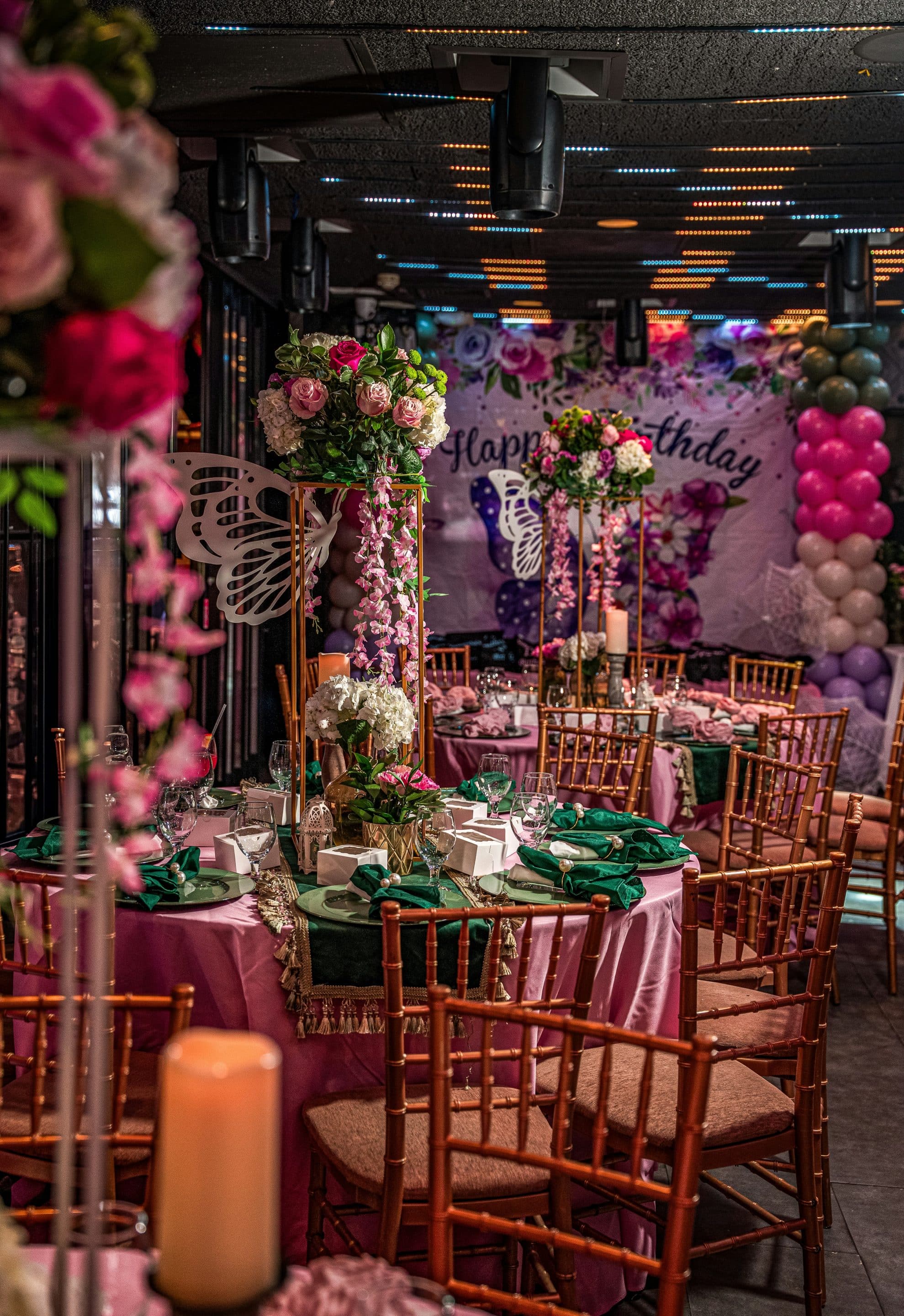 A festive birthday party setup with round tables covered in pink tablecloths, wooden chairs, floral centerpieces, and green napkins. The background features a "Happy Birthday" banner and colorful balloon decorations.