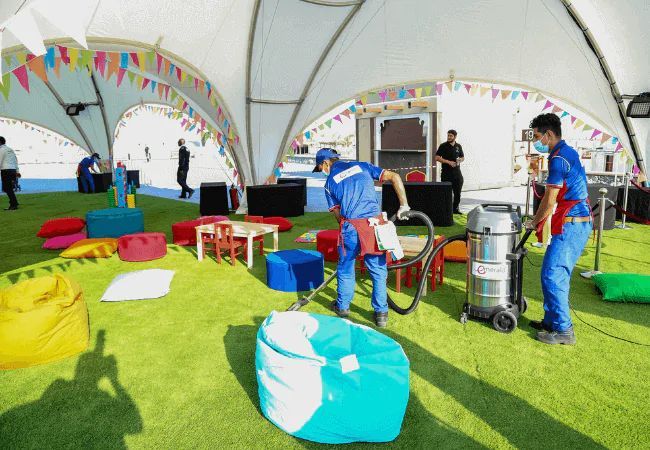 Two workers in blue uniforms clean a grassy indoor event space with bean bags, small tables, and colorful decorations. One uses a vacuum cleaner while the other sprays cleaning solution. Other people are visible in the background.